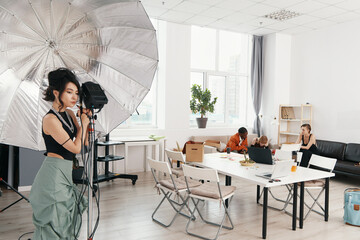 Young woman working with lighting equipment in a modern workspace while colleagues socialize and...
