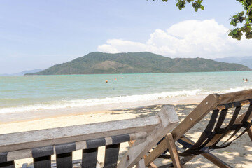 A serene beach with wooden lounge chairs, the sea, a green hill on the horizon, and a clear sky.