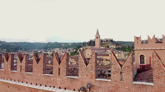 Drone orbits the red-brick crenellated walls of Castelvecchio in Verona, with San Zeno&rsquo;s bell tower and cypress-lined hillside in the background, real time, wide aerial motion, historic urban view