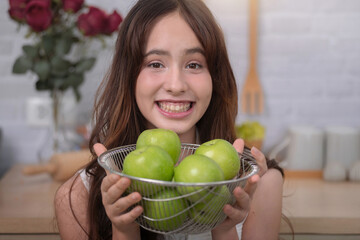 Close up, happy teenage girl holding basket full of green apples in home kitchen, looking positive...