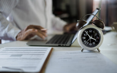 selective focus on alarm clock, asian businessman working with laptop and document on table before...