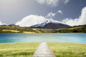 Wooden boardwalk is running through vector grass toward lake with hills, snow-capped peak, clouds
