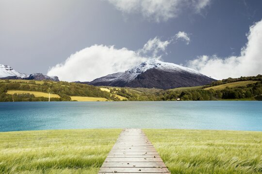 Fototapeta Wooden boardwalk is running through vector grass toward lake with hills, snow-capped peak, clouds