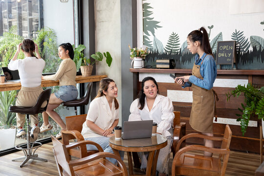Young Asian women working on laptop with coffee cups while talking to female waitress taking order in cozy cafe. Background features two more women relaxing at high table by large window.