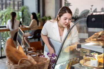 Young Asian businesswoman holding laptop standing near dessert display choosing cake in bakery cafe. Background shows two women relaxing at window counter with plants and natural light.