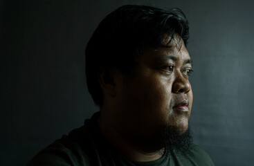 Bearded Indonesian man on black background, neutral expression in low-key lighting, simple portrait...