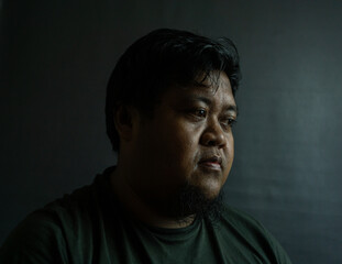 Bearded Indonesian man on black background, neutral expression in low-key lighting, simple portrait...