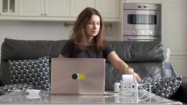 A woman uses her laptop and a label printer at her glass table in her home office, likely managing tasks for her online store.