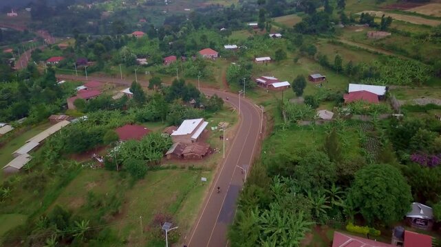 Drone tracking shot of a motorcycle riding along a curved road lined with banana plants and houses near Mount Elgon in Uganda, with fields, trees, and rural buildings forming a lush green landscape