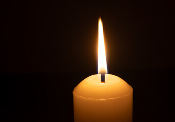 A single burning candle flame or light glowing on a big white candle isolated on black or dark background on table in church for Christmas, funeral or memorial service