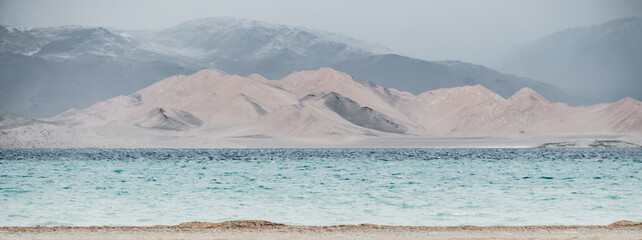 Grass and stone shore of Lake Karakul with blue water and mountain peaks in the background, cloudy...