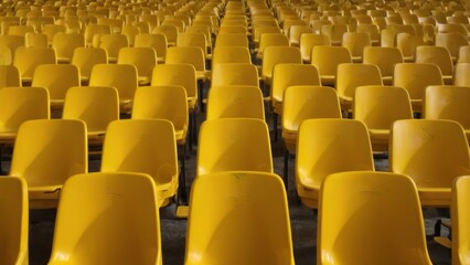 Rows of empty yellow chairs in stadium arranged neatly, facing same direction, stand out against dark background. Emptiness and neglect.