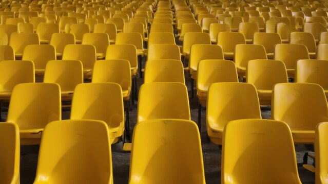 Rows of empty yellow chairs in stadium arranged neatly, facing same direction, stand out against dark background. Emptiness and neglect.