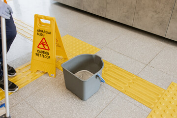 Wet floor caution sign with cleaning bucket and mop in indoor public space, warning pedestrians of...