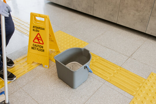 Wet floor caution sign with cleaning bucket and mop in indoor public space, warning pedestrians of slippery surface, placed on tactile paving tiles for visually impaired guidance