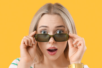 Shocked young beautiful woman in stylish sunglasses on yellow background
