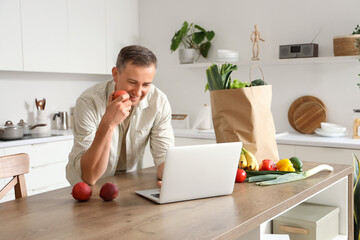 Mature man with apple using laptop at table in kitchen