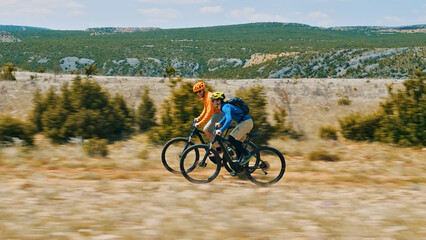 Two helmeted cyclists pedal their e-bikes through a scenic, open field with mountains in the...