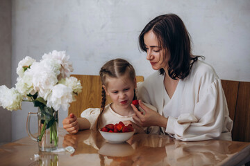 A family of mother and daughter together in the kitchen at the table eating strawberries, beautiful...
