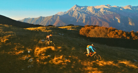 Two mountain bikers traverse a grassy field with a stunning mountain range in the background. This dynamic aerial view showcases their thrilling outdoor adventure. © wkproduction