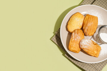Plate with delicious madeleines, sieve and powdered sugar on green background