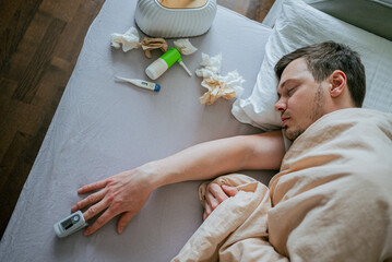 Sick man is sleeping in bed with a pulse oximeter on his finger, surrounded by tissues, nasal...