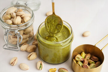 Jar of pistachio paste with spoon and measuring cup on white background
