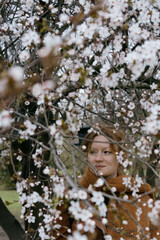 woman with red hair stands in front of a tree with pink flowers. She's wearing a brown coat, scarf.