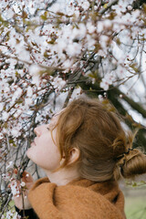 woman with red hair stands in front of a tree with pink flowers. She's wearing a brown coat, scarf.