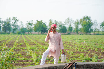 young indian farmer standing at field talking on smartphone