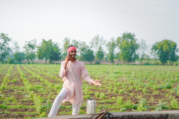young indian farmer standing at field talking on smartphone