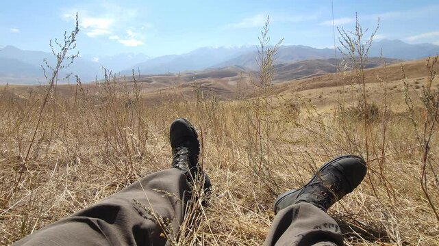 First-person perspective POV shot of legs in military combat boots resting in dry grass, enjoying the mountain landscape view in Kyrgyzstan.