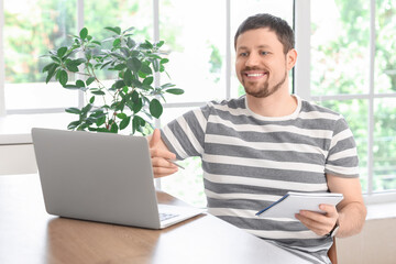 Young male teacher with laptop and notebook giving online lesson at home