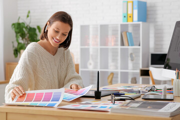 Young female designer working with graphics tablet and color samples at table in office