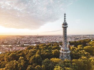 Obraz premium Golden light bathes Petrin Lookout Tower as the sun rises over Prague. Surrounded by lush greenery and the city skyline, this moment captures the beauty of early morning tranquility.
