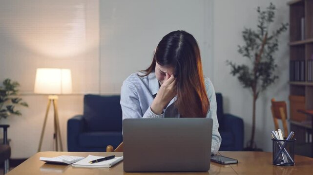 Professional young Asian businesswoman experiencing workplace stress while working remotely on laptop computer, revealing mental health challenges in home office environment