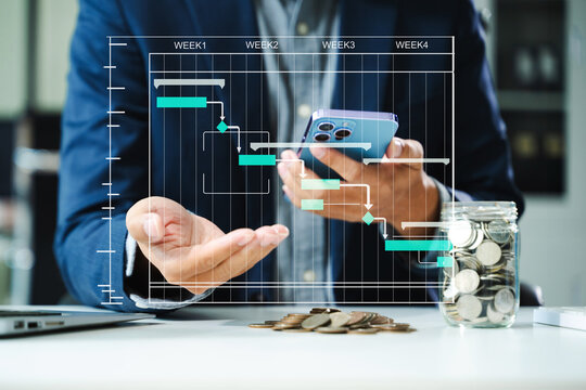 Project planning and finance concept with Gantt chart, coins, and smartphone in office. Businessman visualizes productivity