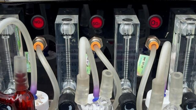 Laboratory gas analysis system with labeled flow meters for NO₂, SO₂, and O₃, transparent tubing, and chemical absorption bottles used for environmental air quality testing. Impinger machine.
