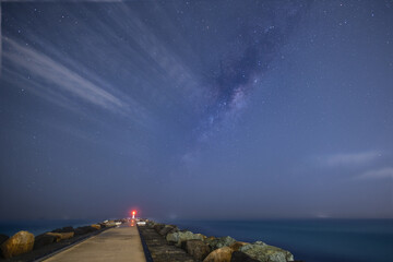 A long exposure, astrophotography view of the milky way stars, and clouds above the red navigation light at the end of the breakwater jetty at The Spit on the Gold Coast in Queensland, Australia.