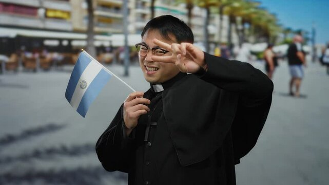 Asian man priest smiling holding salvadoran flag on busy urban street with buildings and people in background.