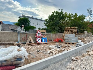 Pile of road signs, cement bags, pallets, and construction materials on a sidewalk next to a fence....