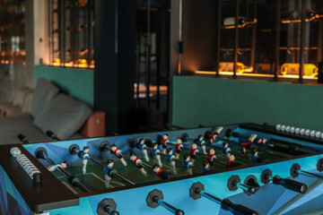 Foosball table with vibrant blue sides and miniature players sits in a stylish lounge bar; backlit...