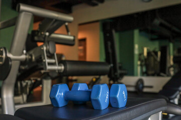 Pair of small blue hex dumbbells resting on a workout bench in a modern, empty gym; strength-training equipment blurred in the background