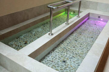 Stone-lined Kneipp footbath with stainless rail and green-purple LED lights in a modern indoor spa...