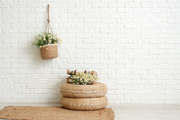 Suitcase with chamomiles on rattan poufs near white brick wall in room
