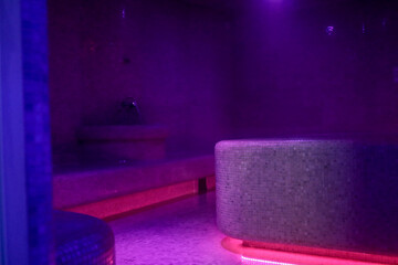 Violet-lit steam room with misty air, mosaic-tiled benches and a glowing heated centerpiece create...