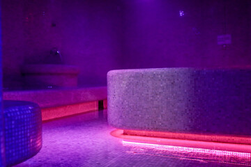Violet-lit steam room with misty air, mosaic-tiled benches and a glowing heated centerpiece create...