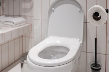 Clean modern hotel toilet with white ceramic bowl, open seat, wall-mounted paper roll and tiled...