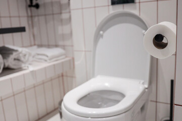 Clean modern hotel toilet with white ceramic bowl, open seat, wall-mounted paper roll and tiled...