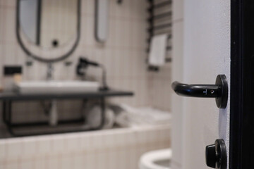 Close-focus shot of black door handle with modern hotel bathroom softly blurred behind—oval mirror, washbasin and towels—conveying clean contemporary design